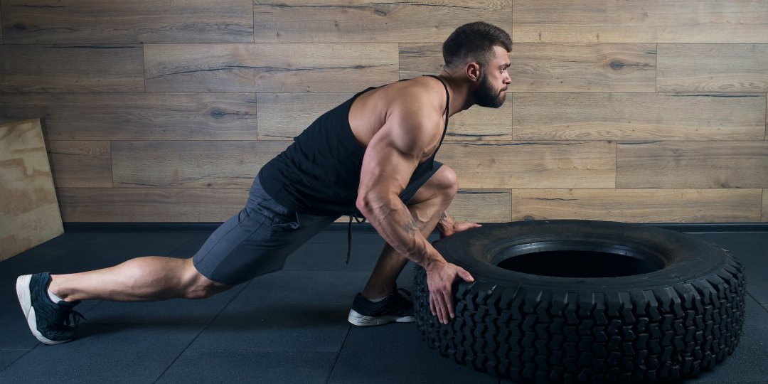 A bodybuilder with a tattoo and beard is pushing a tire in a gym